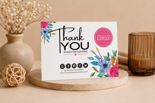 A printed thank-you card with floral artwork and black text reading 'Thank You So Much for Your Order,' styled on a round stone coaster with a beige vase, dried flowers, and an amber glass candle holder in the background.